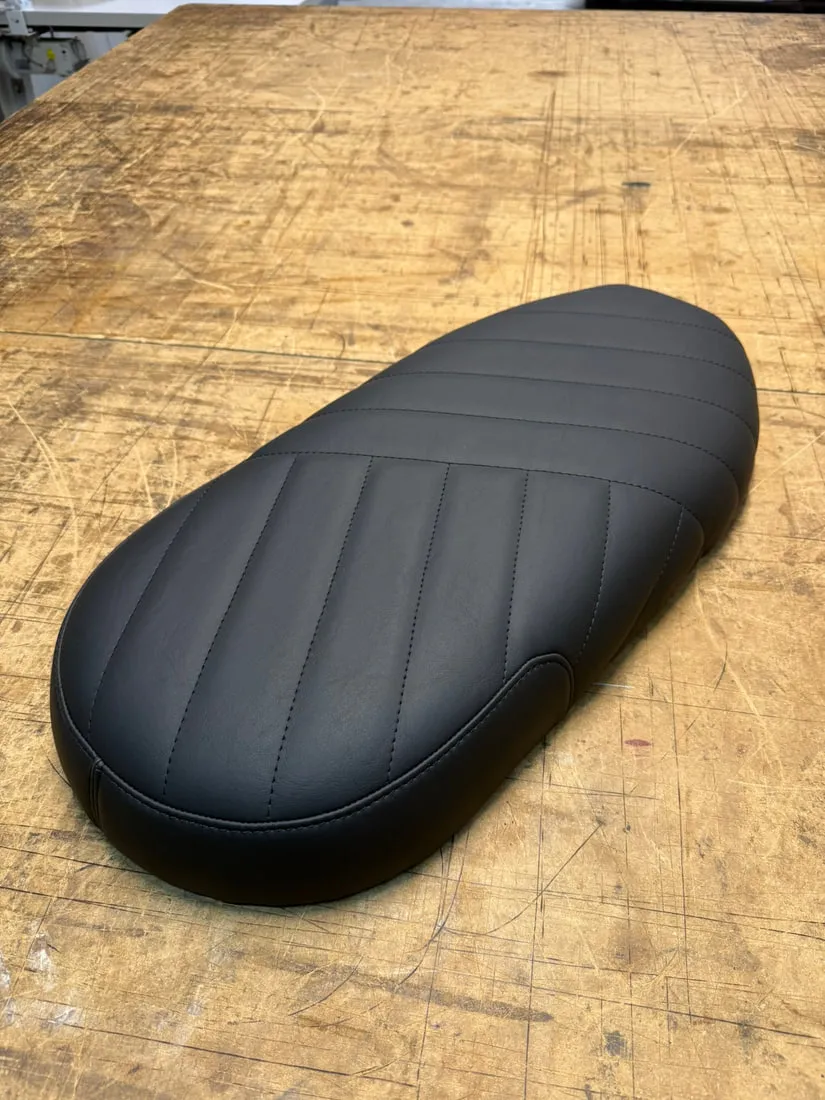 Selle de Triumph Bonneville refaite en simili noir mat à surpiqûres longitudinales, posée sur l’établi